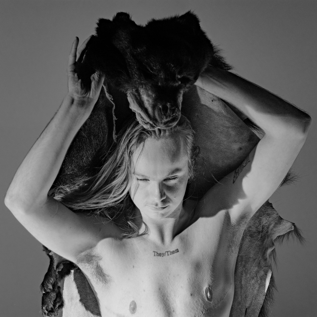 A shirtless person looks downward and holds an American black bear skin above their head. The words "they/them" are tattooed on their upper chest. Top surgery scars are visible on their lower chest.