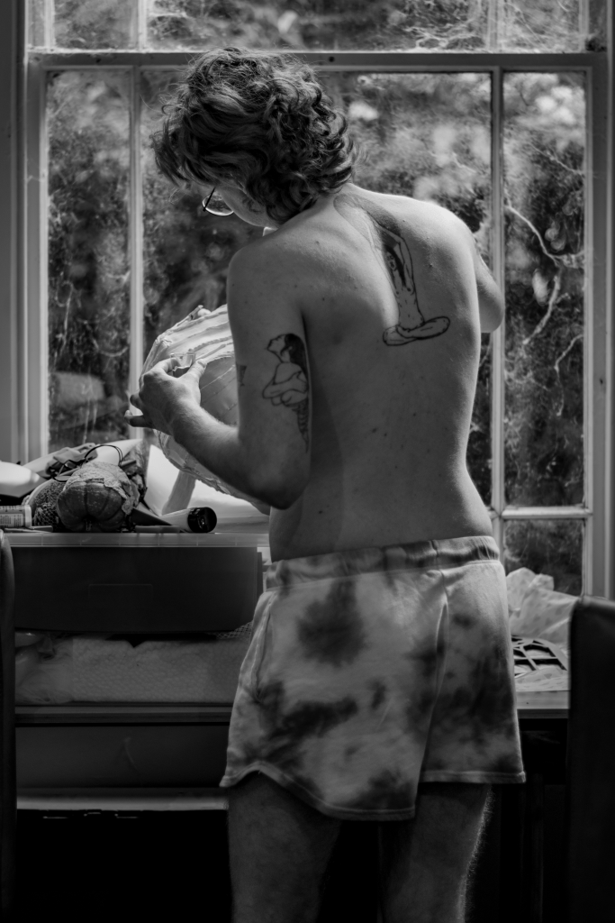 A shirtless person stands with their back to the camera in front of a window, looking at and holding something unseen. 
