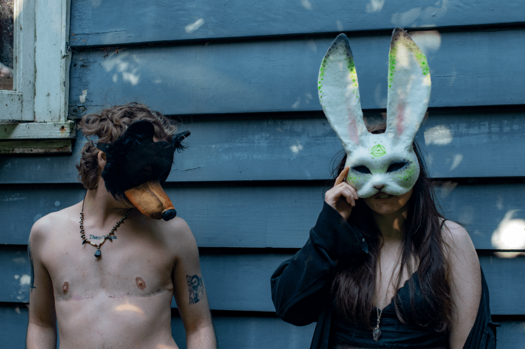 Two people in homemade therian masks stand next to each other in front of an old house. The one on the left wears a bear mask and the one on the right wears a rabbit mask.